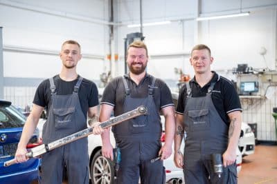Drei BMW-Service-Mechaniker im grauen Arbeitsoverall stehen lächelnd in einer Werkstatt; einer hält einen langen Drehmomentschlüssel, ein anderer eine Akku-Bohrmaschine, der Dritte einen Schlagschrauber.