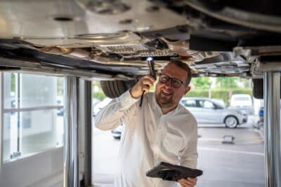 Serviceberater von Kattenstroth Automobile inspiziert unter einem aufgebockten Fahrzeug den Unterboden: Er hält eine Taschenlampe in der einen und ein Tablet in der anderen Hand, lächelnd in Richtung Kamera.