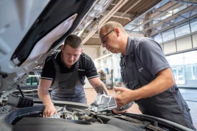 Zwei BMW-Servicetechniker von Kattenstroth Automobile prüfen unter geöffneter Motorhaube ein Trägerteil: Der ältere Monteur hält ein Bauteil in der Hand, der jüngere tastet am Motorblock, beide konzentriert im hellen Werkstattbereich.