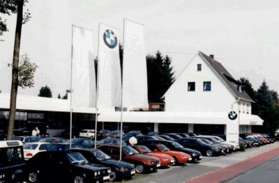 Historische Fotografie des BMW Autohauses Kattenstroth: Viele ältere 3er-Limousinen und Coupés stehen vor einem weißen Verkaufsgebäude mit Fahnenmast und Einfamilienhaus-Dachaufbau.
