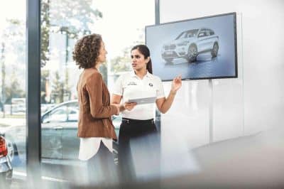 Verkaufsberatung im BMW-Showroom: Eine BMW-Mitarbeiterin im weißen Poloshirt zeigt einer Kundin mit Tablet die Features eines SUV, im Hintergrund ein Bildschirm mit Fahrzeuggrafik.