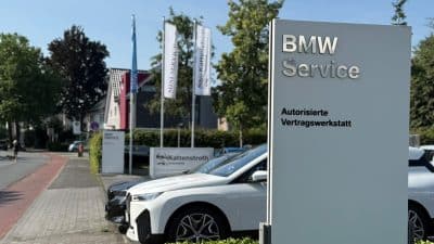Außenansicht der Vertragswerkstatt: Ein hoher totemartiger Pylon mit Schriftzug „BMW Service – Autorisierte Vertragswerkstatt“, dahinter Fahnen und geparkte Wagen am Straßenrand, der Fahrzeugbestand von Kattenstroth Automobile in Gütersloh.