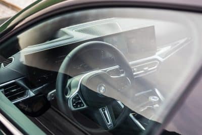 Blick durch die Windschutzscheibe auf das Cockpit eines BMW: Lenkrad mit blau-schwarzem BMW-Emblem, großformatiges Kombiinstrument und Infotainment-Display im Armaturenbrett.
